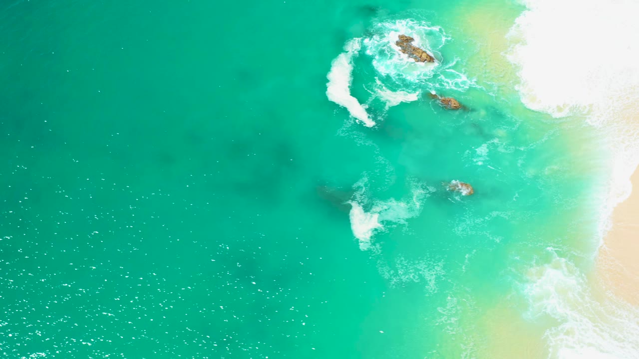 White foam curls around rocks in clear turquoise water near sand.