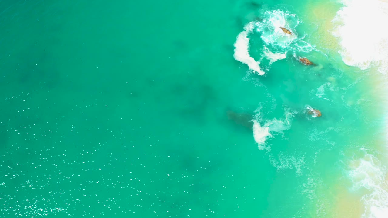 Aerial view over open teal water at Marquis Los Cabos, Mexico, with wave foam and rocks concentrated near the upper right, a pale sandy edge, and no visible people.