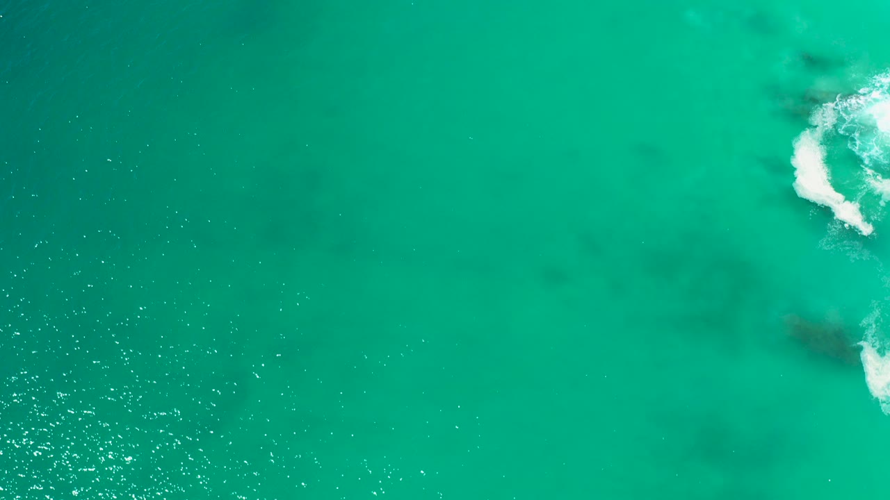 Overhead view of turquoise ocean at Marquis Los Cabos, Mexico, with dark submerged patches, sun flecks, white surf near the right side, and no visible people.