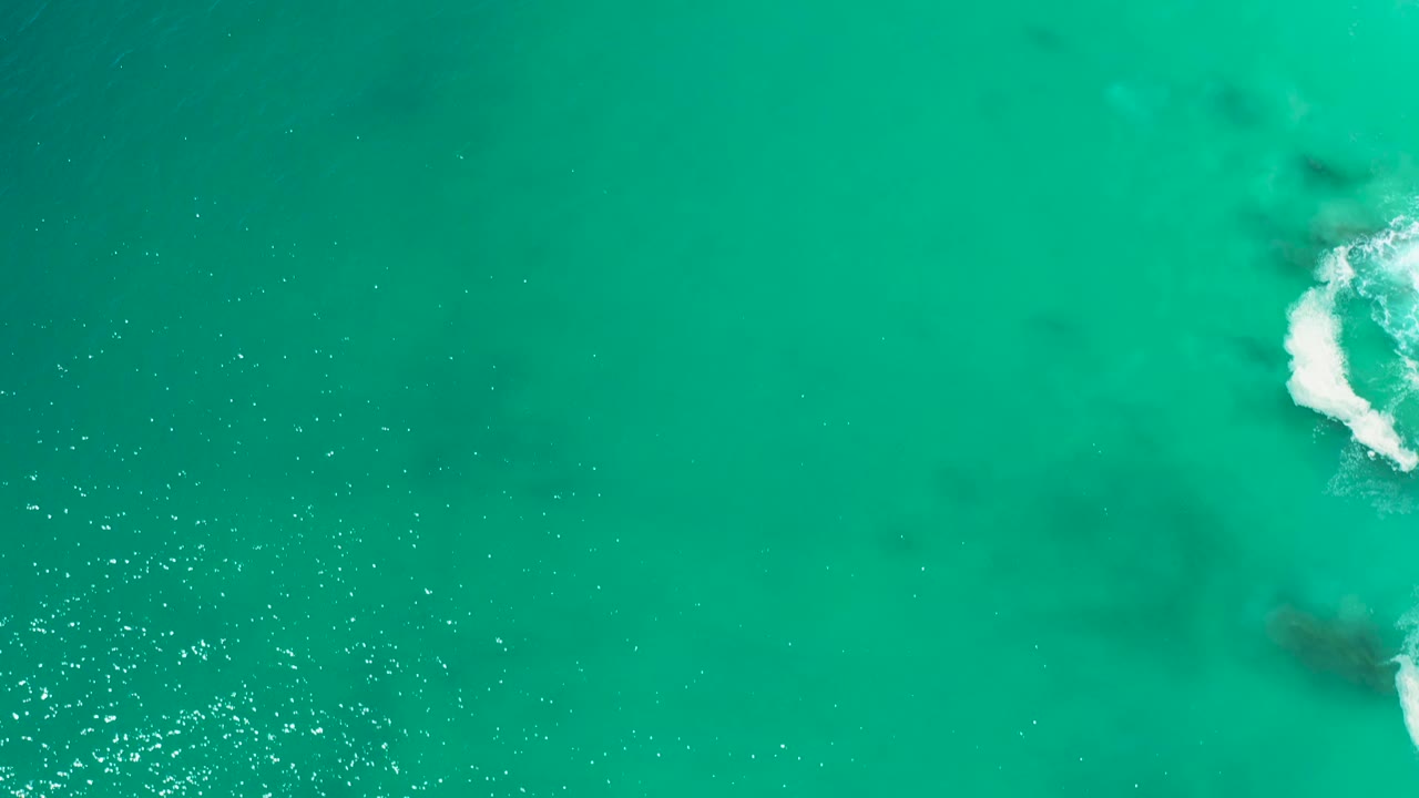 Top-down view of turquoise sea at Marquis Los Cabos, Mexico, with white surf partly cropped on the far right, faint dark patches below the water, and no visible people.