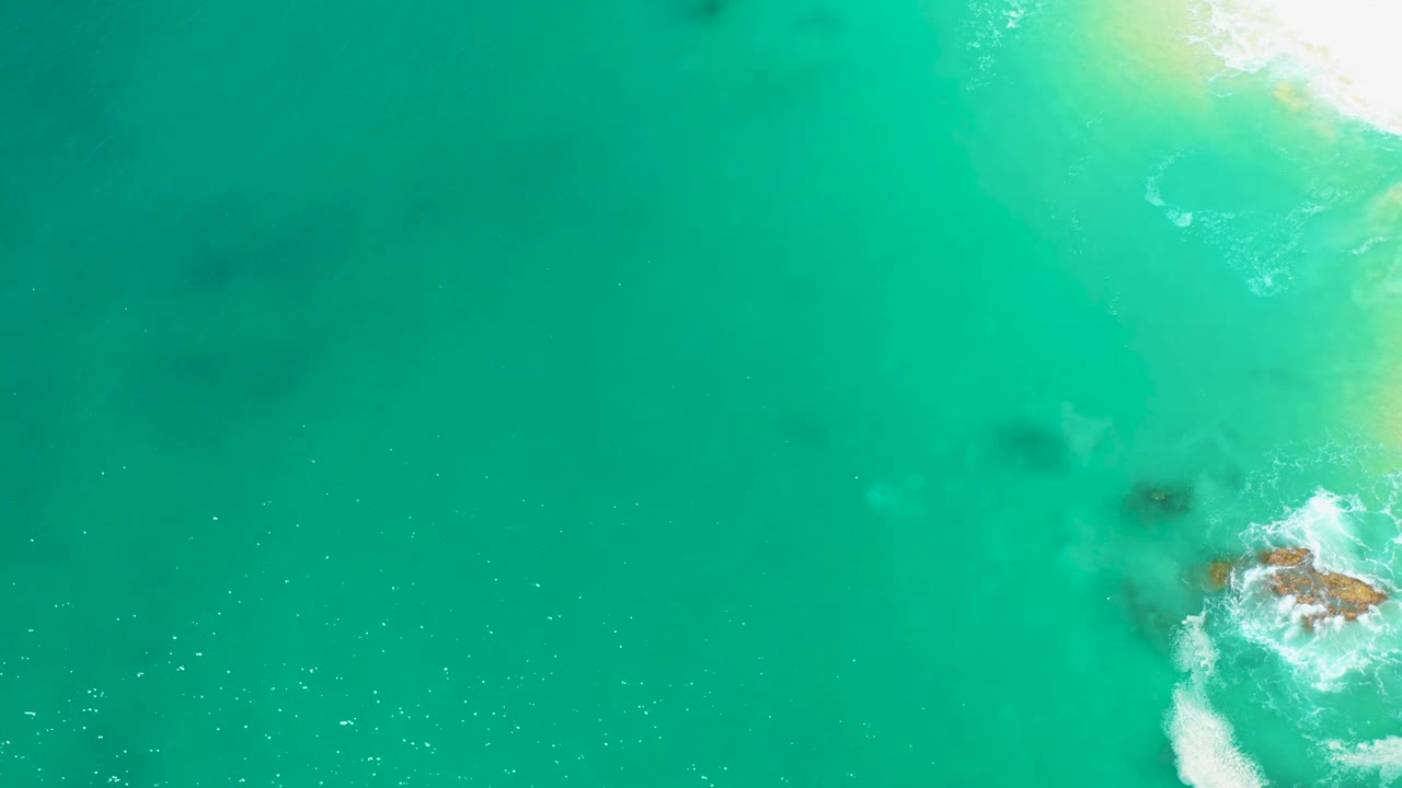 Aerial frame from Marquis Los Cabos with open turquoise water, a foamy wave curling around a small rock near the sandy shore, and no visible people.