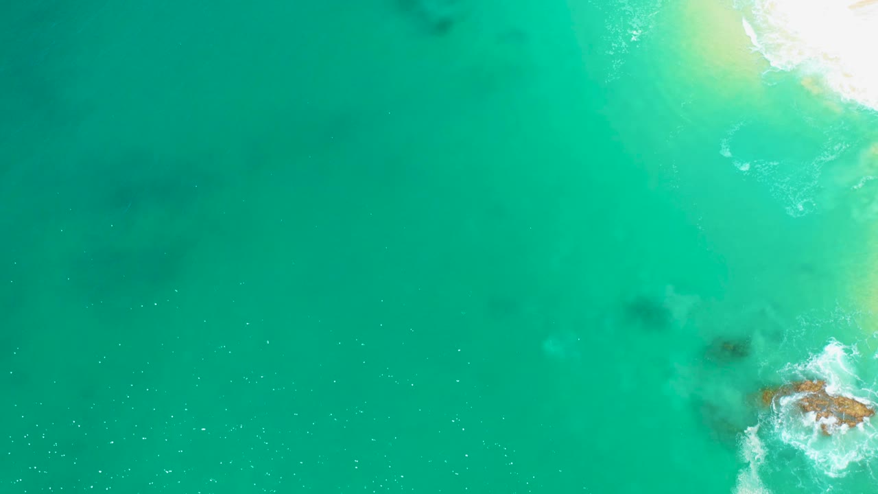 Top-down Marquis Los Cabos shoreline view with broad turquoise water, pale sand and white foam along the right side, faint underwater shadows, and no visible people.