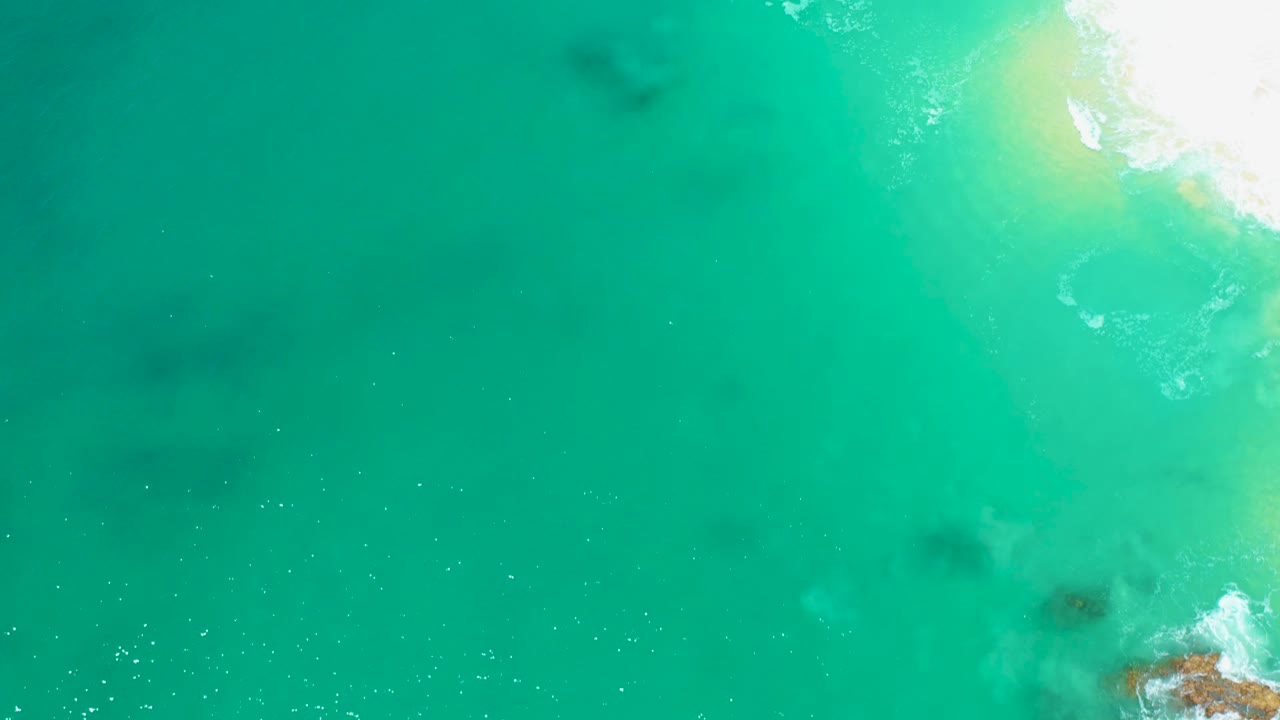 Overhead Marquis Los Cabos frame with mostly turquoise water, bright foam and pale sand on the right edge, darker water patches, and no visible people.