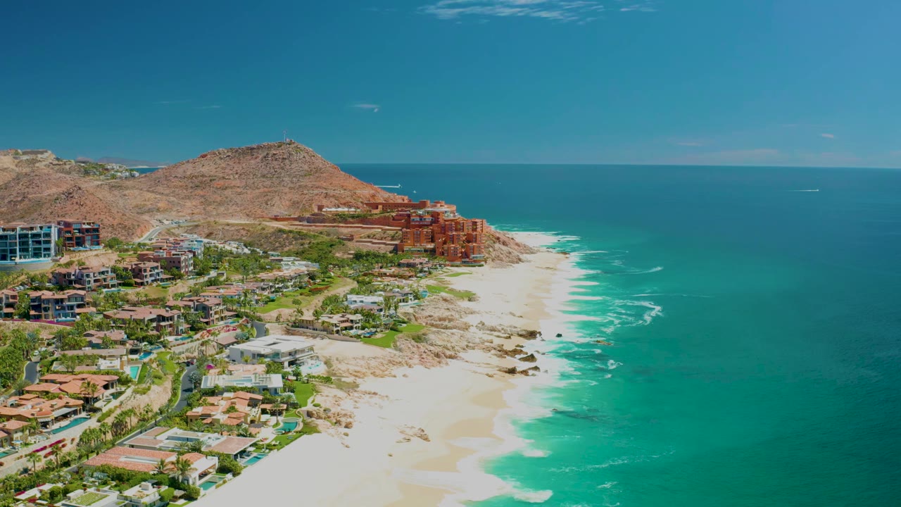 Aerial view at 0:01 over Marquis Los Cabos, Mexico, with surf spreading along the beach and the hillside buildings slightly more centered, no visible people.