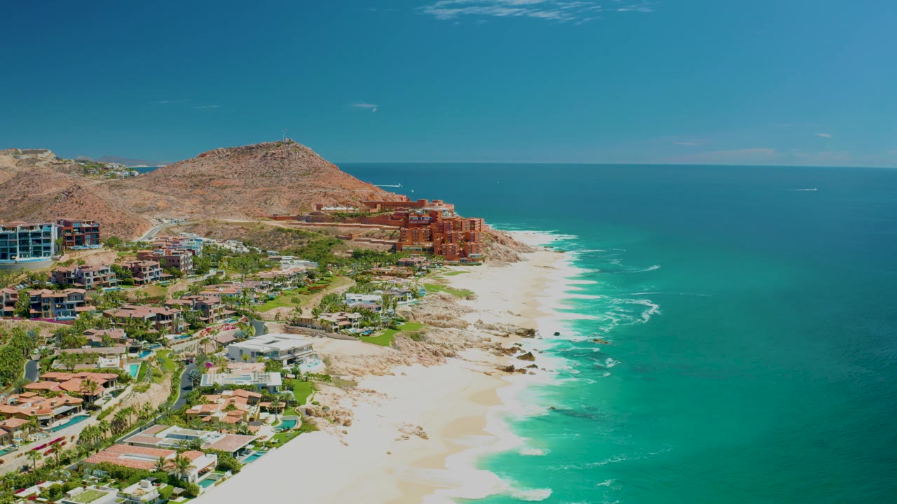 At 0:02, the Marquis Los Cabos coastline remains in a wide aerial composition, with bright surf marks on the sand and no visible people.