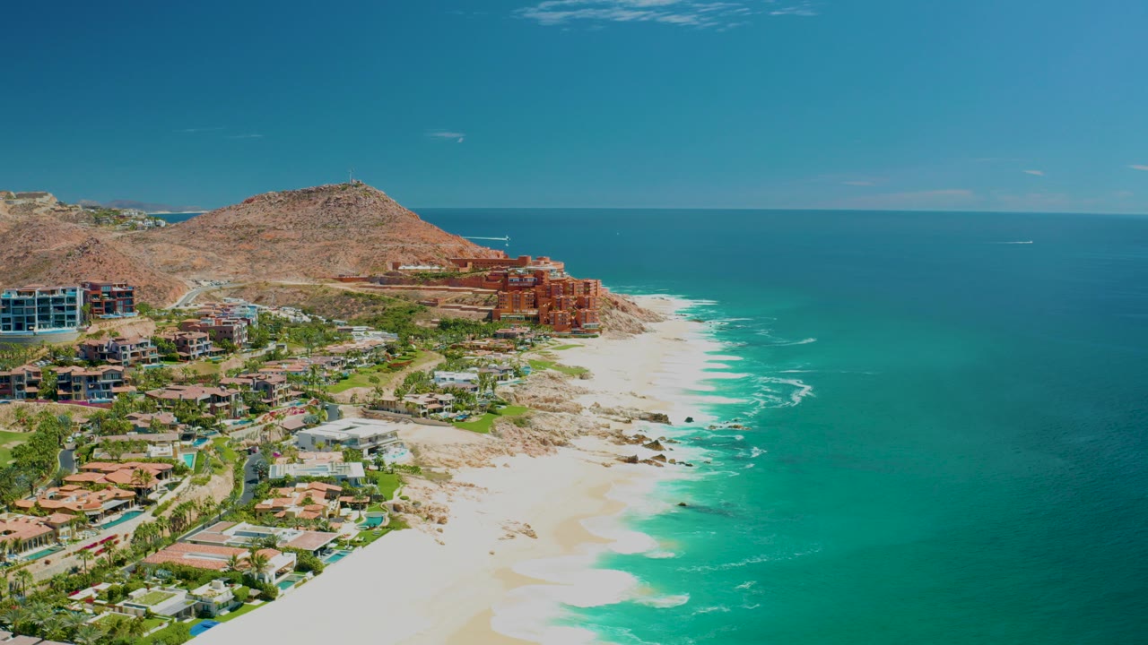 Opening aerial frame of Marquis Los Cabos with hillside buildings on the left, a bright sandy beach below, turquoise ocean to the right, and no visible people.