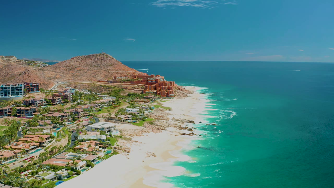 Aerial view of Marquis Los Cabos showing more of the resort buildings and hillside at left, a pale beach curving along the surf, and no visible people.