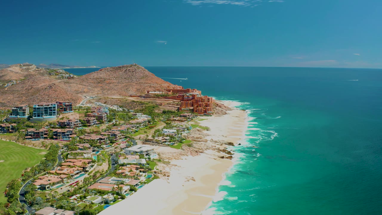 Aerial view at Marquis Los Cabos, Mexico with the resort buildings centered lower left, white surf tracing the sandy shoreline, open blue water on the right, and no visible people.