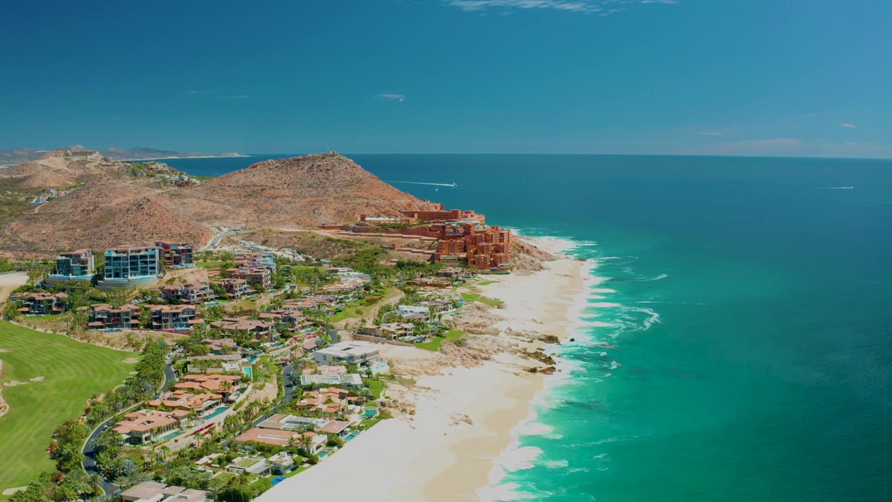 Aerial frame from Marquis Los Cabos, Mexico with green landscaped grounds at lower left, resort buildings near the sand, waves along the shoreline, and no visible people.