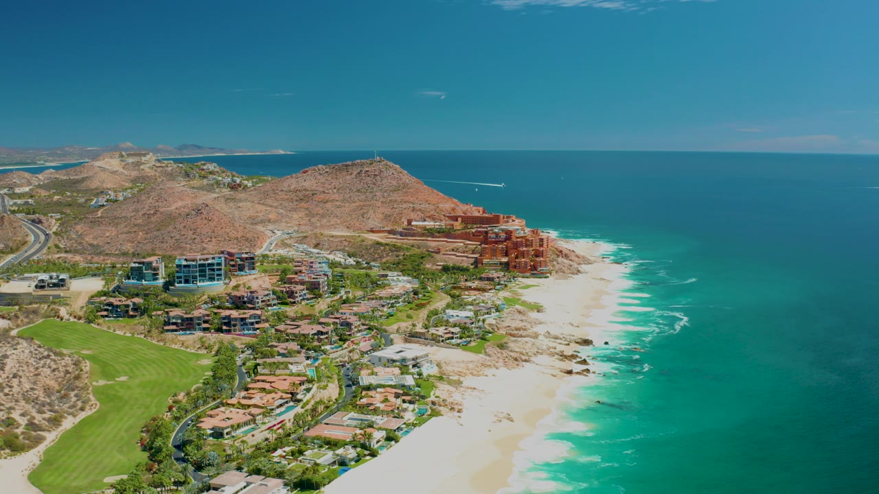 Opening aerial frame over Marquis Los Cabos, Mexico, showing a coastal road at left, green grounds, sandy beach, hillside buildings, turquoise ocean, and no visible people.