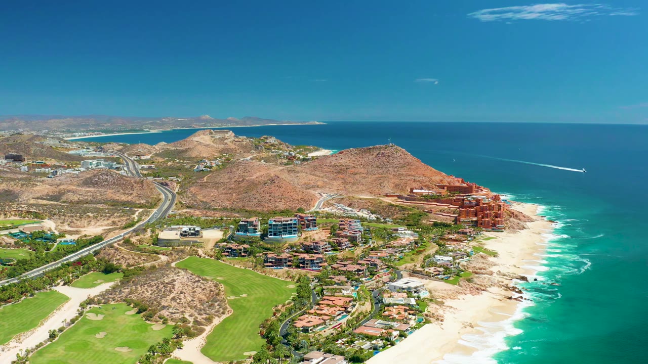 Opening aerial frame over Marquis Los Cabos, Mexico, showing a sandy beach, turquoise surf, hillside buildings, a coastal highway, green grounds, and no visible people.