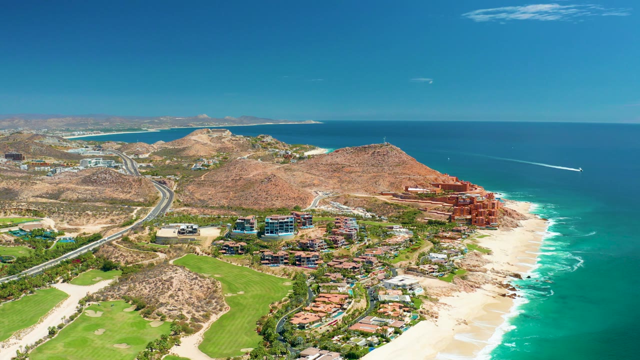 Mid-clip aerial frame over Marquis Los Cabos, Mexico, with green grounds in the lower left, the highway on the hillside, beach and surf on the right, and no visible people.