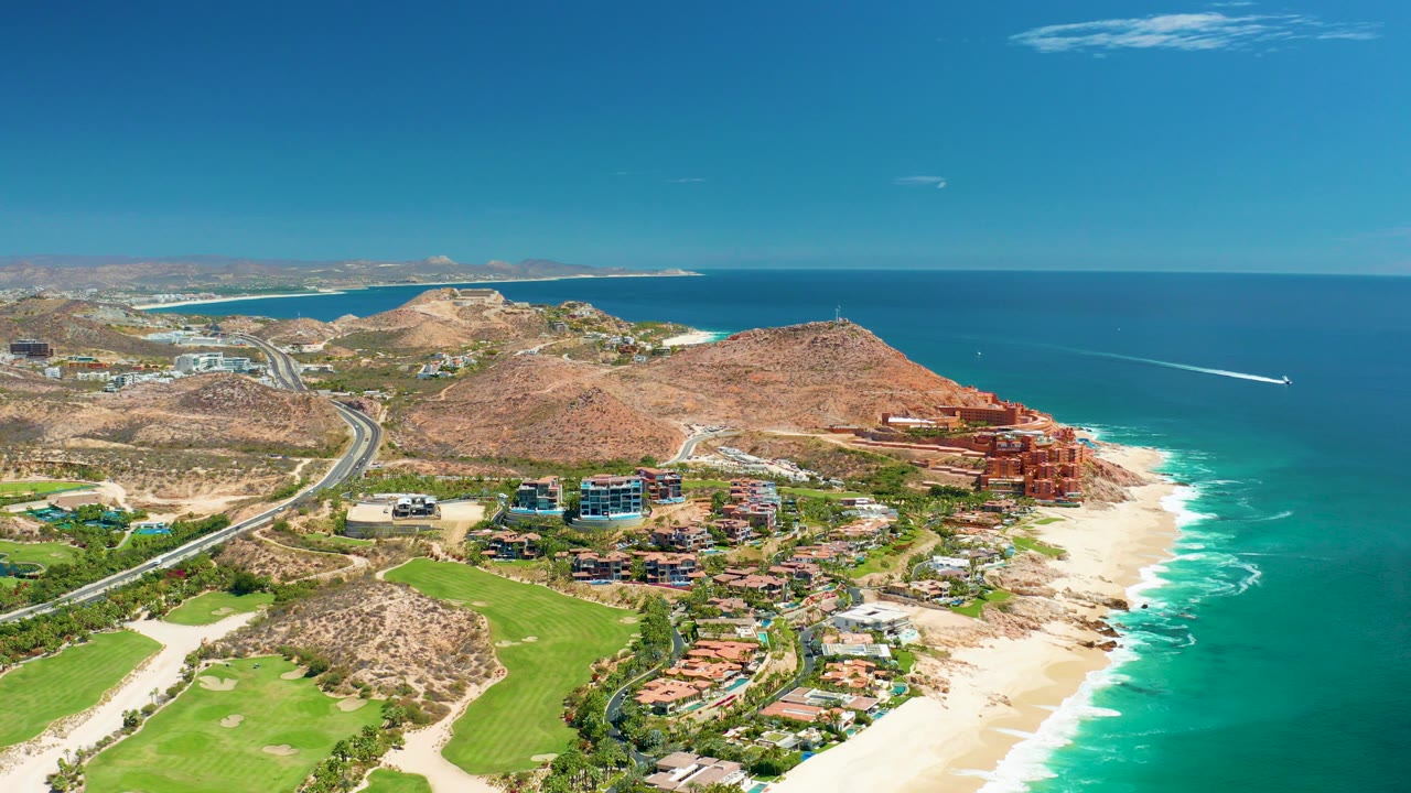 Opening aerial frame over Marquis Los Cabos, Mexico with a coastal highway, sandy beach, turquoise ocean, arid hills, green landscaped ground, resort-area buildings, and no visible people.