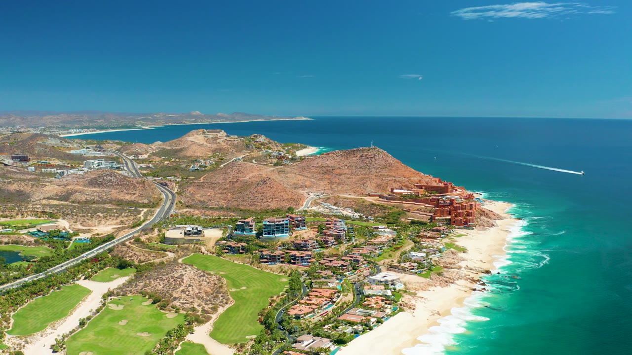Opening aerial frame over Marquis Los Cabos, Mexico, showing a coastal highway, green fairways, sandy beach, resort buildings, blue ocean, and no visible people.