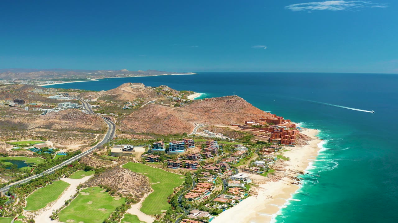 Wide aerial frame over Marquis Los Cabos showing golf greens in the lower left, a curving coastal road, dry hills, red coastal buildings, sandy beach, turquoise surf, open ocean, and no visible people.