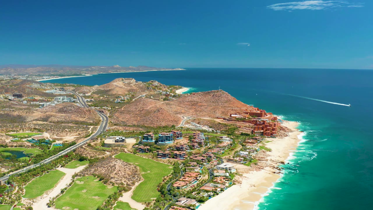 High coastal aerial with road, hills, buildings, beach, ocean, and a boat wake offshore.
