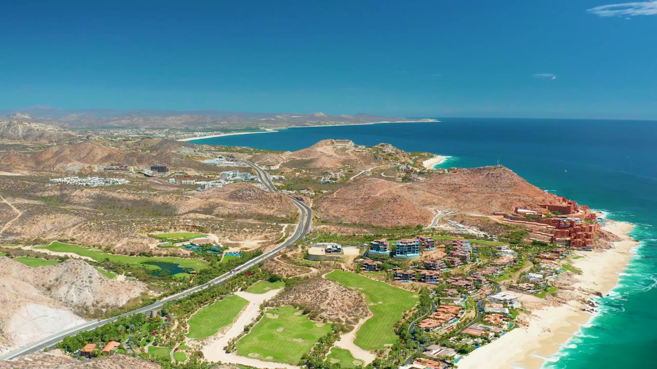 Wide aerial frame over Marquis Los Cabos, Mexico showing a highway beside green golf fairways, sandy beach, turquoise surf, dry hills, and blue ocean, with no visible people.