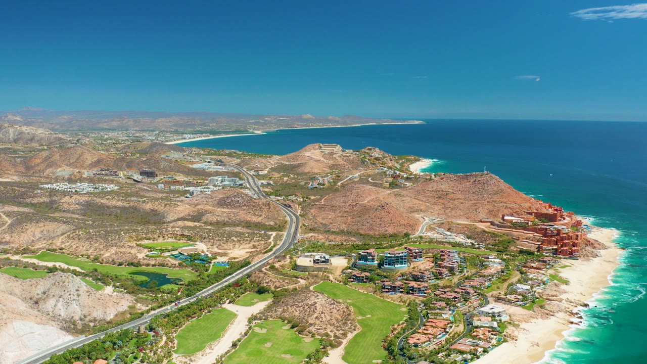 Opening aerial frame over Marquis Los Cabos, Mexico, showing a coastal highway beside green land, sandy beach, turquoise surf, arid hills, and blue ocean with no visible people.