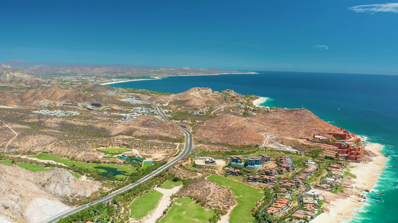 A wide aerial view in Marquis Los Cabos, Mexico, frames the curving highway, dry hills, turquoise shoreline, green grounds, clustered coastal buildings, and no visible people.