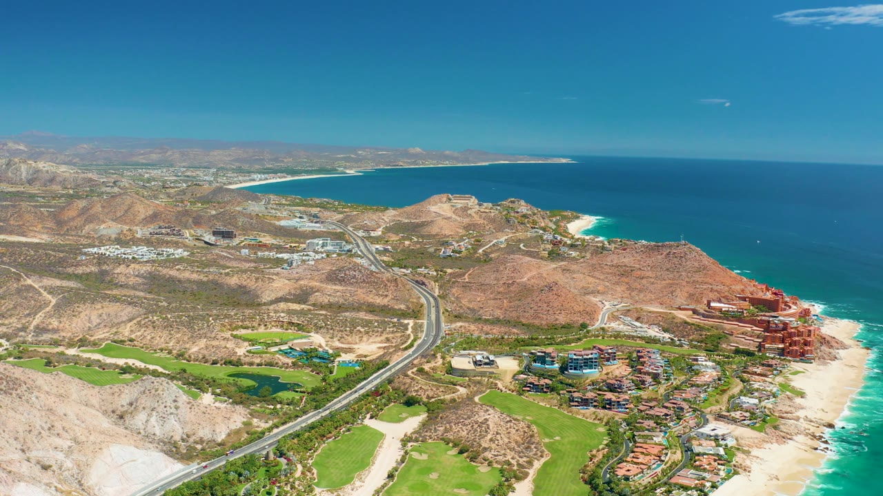 Aerial coastline view with road, hills, beach, ocean, green areas, and buildings near shore.