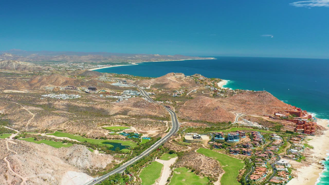 Wide aerial frame over Marquis Los Cabos, Mexico showing a coastal highway through rocky hills, green golf course areas, sandy beach, blue ocean, shoreline buildings, and no visible people.