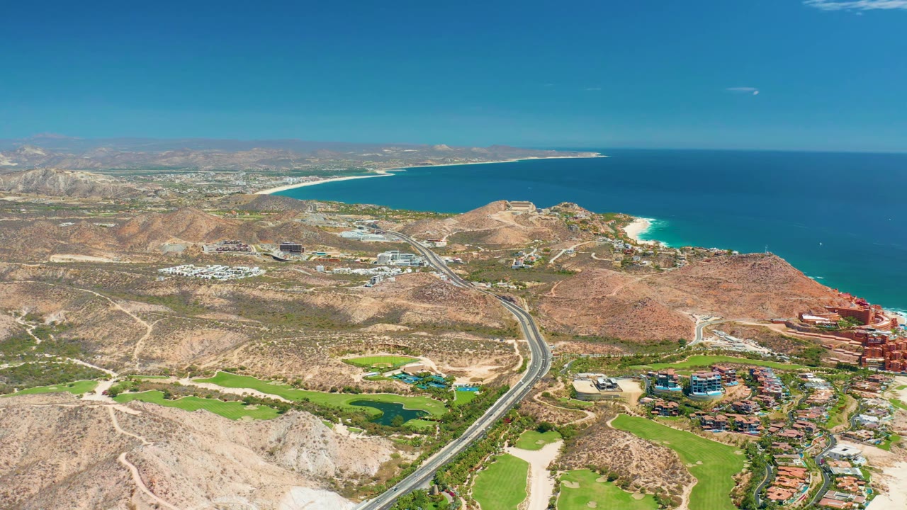Aerial frame from Marquis Los Cabos, Mexico, showing a divided road through tan terrain, green patches below, red and light-colored buildings at the right edge, blue ocean beyond, and no visible people.