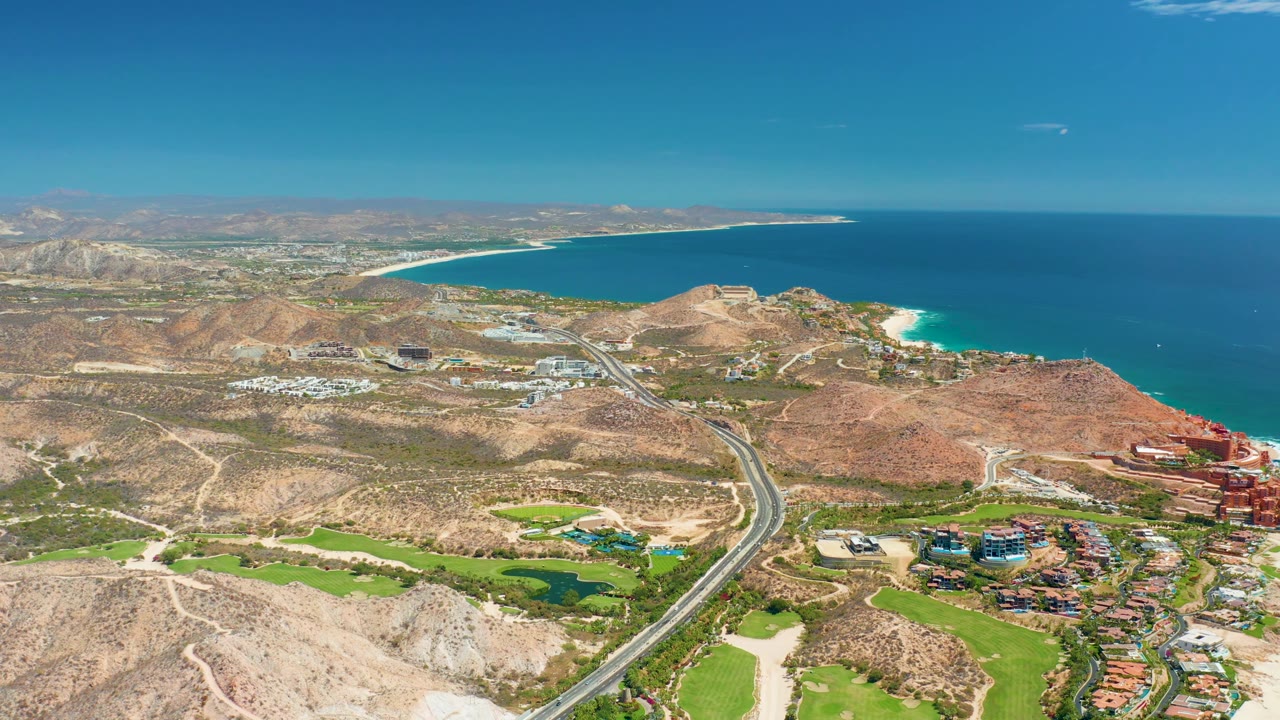 Wide aerial view in Marquis Los Cabos, Mexico, with the curving road centered through dry hills, a turquoise surf edge on the right, developed areas below, broad blue water, and no visible people.