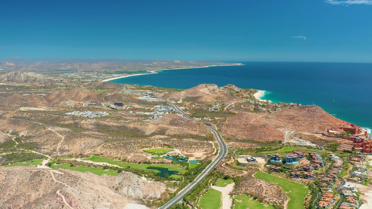 Aerial coastline with winding highway, hills, green landscaping, buildings, and ocean horizon.