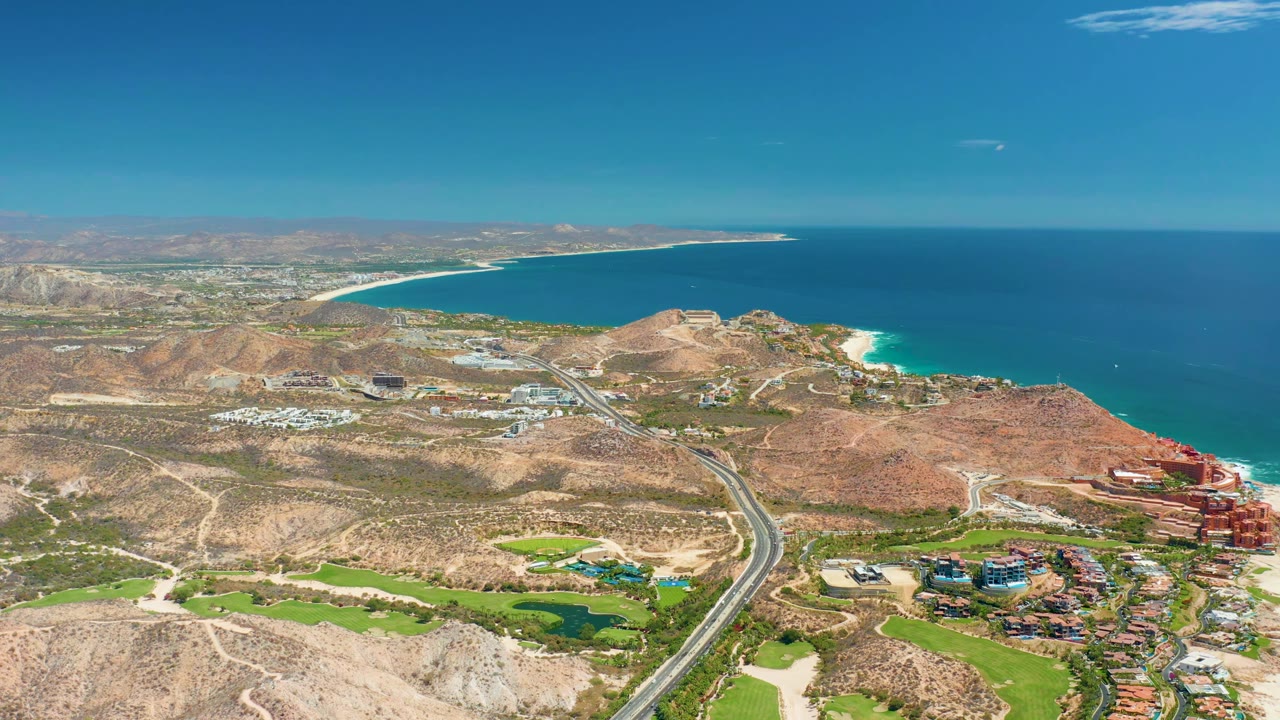 High aerial view at Marquis Los Cabos BCS Mexico showing the highway and dry brown terrain leading toward a curving beach and deep blue ocean, with no visible people.
