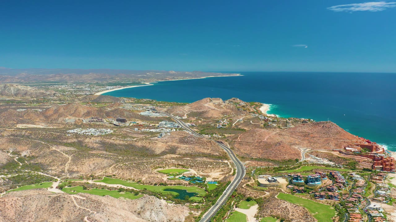 Aerial view in Marquis Los Cabos BCS Mexico with a highway running through arid land, bright green landscaped grounds below, sandy beach beyond, and blue sea under clear sky, with no visible people.