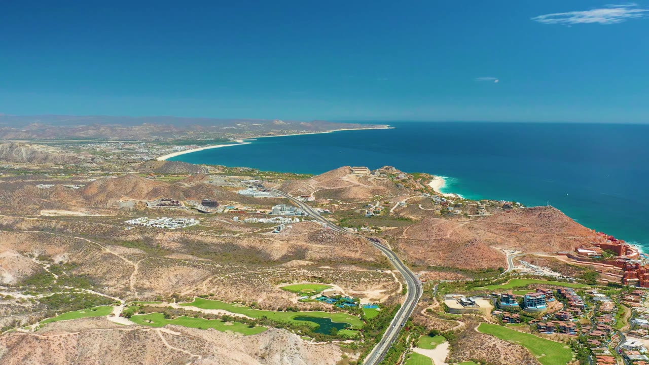 Opening aerial frame over Marquis Los Cabos shows rocky desert hills, a highway, green golf course areas, a sandy coastline, blue ocean, distant mountains, clear sky, and no visible people.