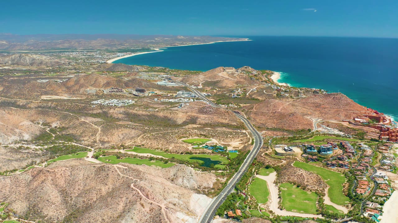 Drone view of coastal highway, golf greens, tan hills, and blue ocean.