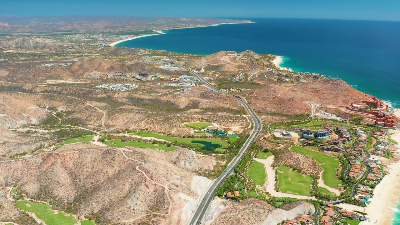 Aerial opening frame over Marquis Los Cabos, Mexico, with a highway running through dry hills toward coastal buildings, a sandy beach, and blue ocean, with no visible people.