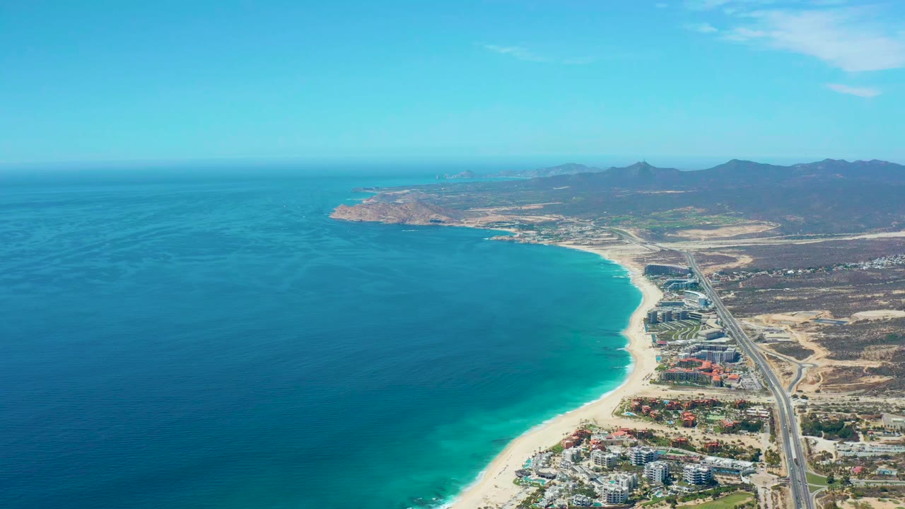 At 0:01, the Marquis Los Cabos aerial keeps the turquoise shallows and pale beach centered, with the highway tracing the coast beside buildings and no visible people.
