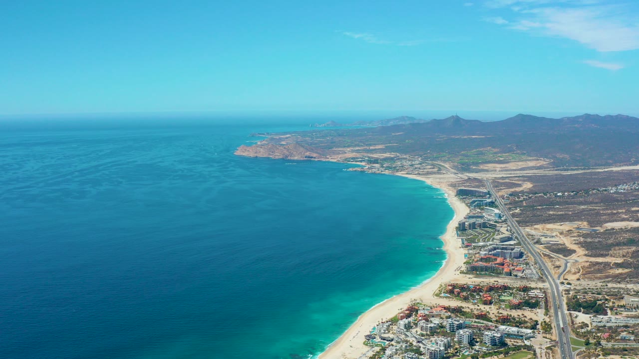Opening aerial frame over Marquis Los Cabos, Mexico, showing turquoise ocean on the left, a curving sandy beach, a parallel highway, coastal buildings, distant mountains, clear sky, and no visible people.