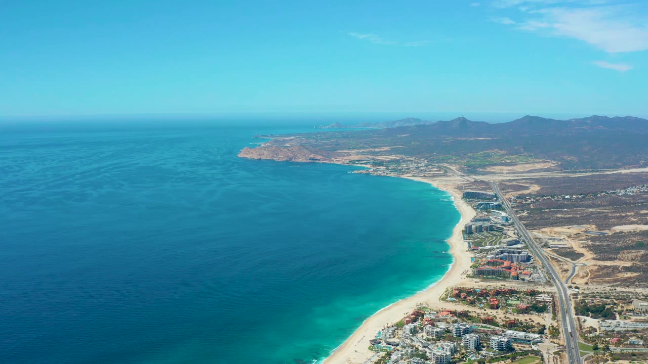 Early aerial frame at Marquis Los Cabos, Mexico, with broad blue ocean water, pale beach, a road running along the shore, clustered buildings near the lower edge, mountains in the distance, and no visible people.