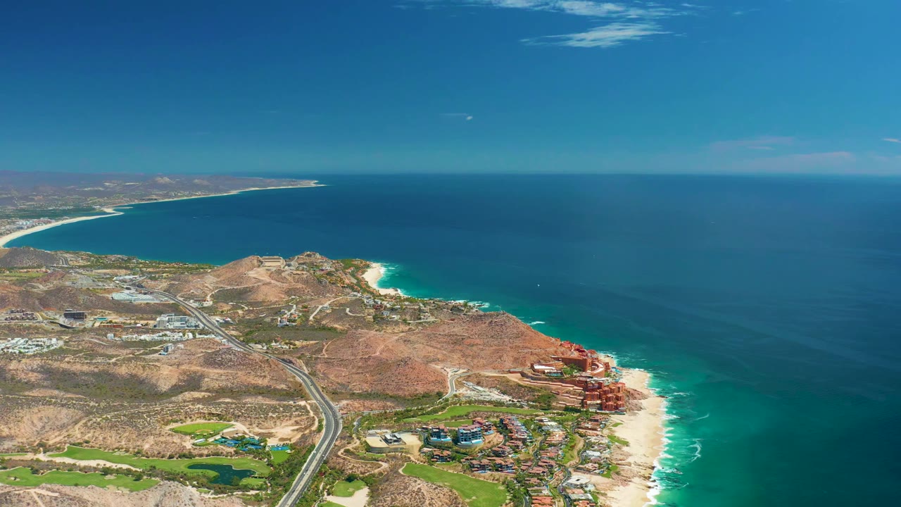 Aerial view of Marquis Los Cabos, Mexico with the shoreline angled through frame, bright turquoise surf at the sand, hillside roads, coastal development, and no visible people at 0:01.
