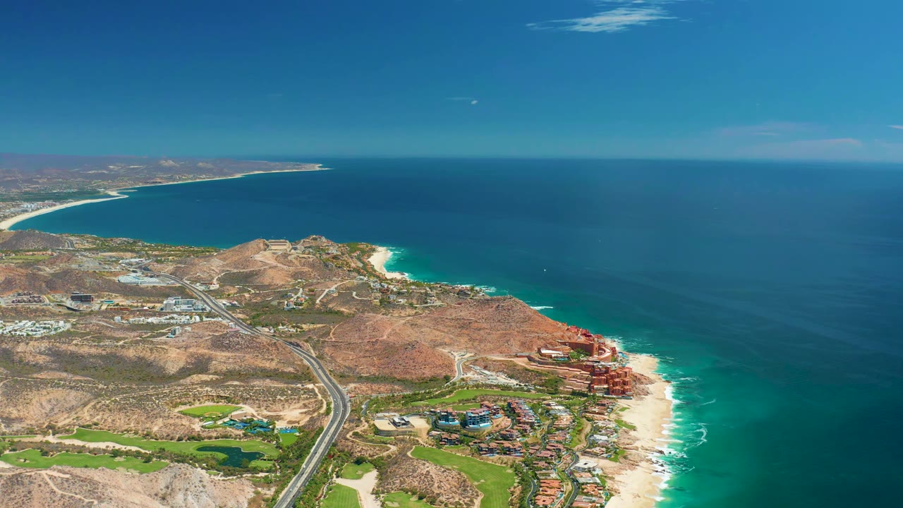 Aerial frame from Marquis Los Cabos showing dry hills, a paved road, resort-area buildings, white surf along the beach, and a small aircraft near the upper sky, with no visible people.