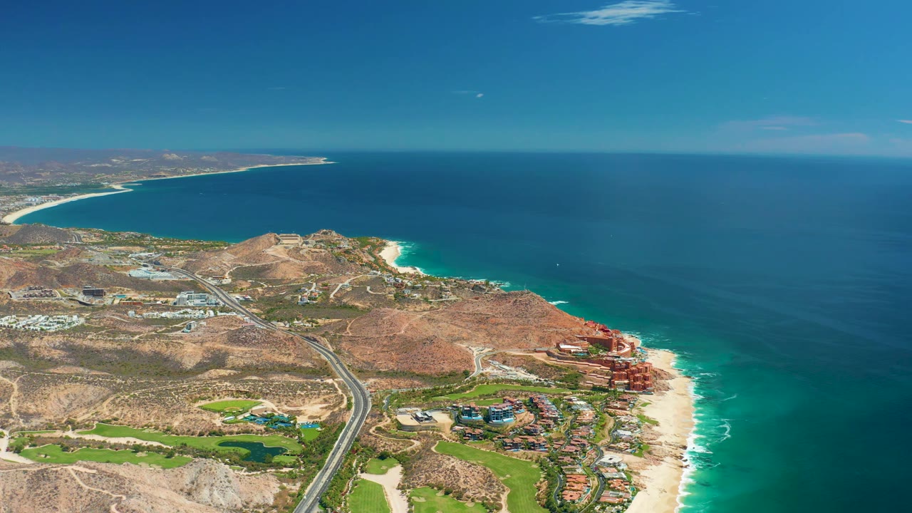High aerial frame over Marquis Los Cabos showing the ocean taking more of the right side, the beach curving below, the road crossing the hills, and no visible people.