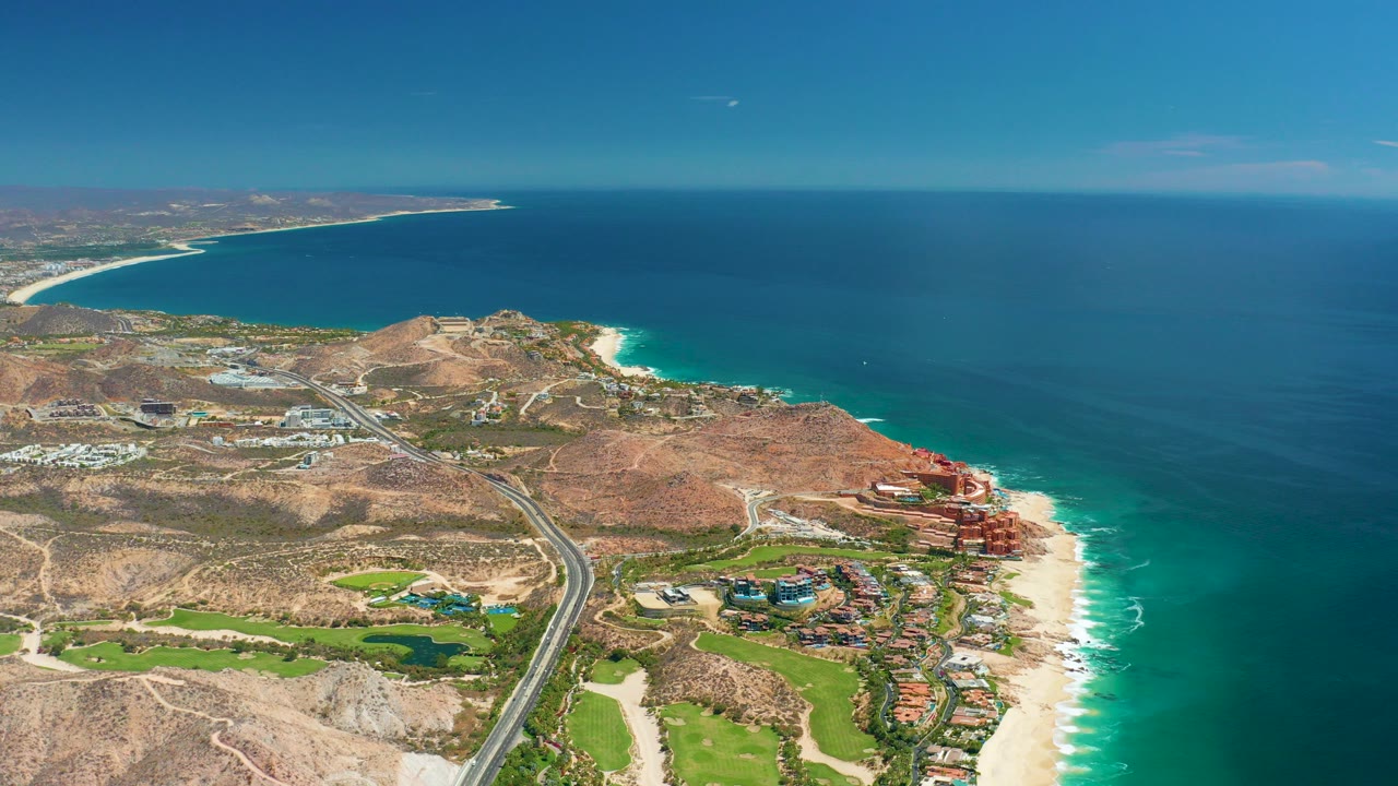 Wide aerial view over Marquis Los Cabos, Mexico, showing a coastal highway through arid hills, green grounds, sandy beach, blue ocean, and no visible people.