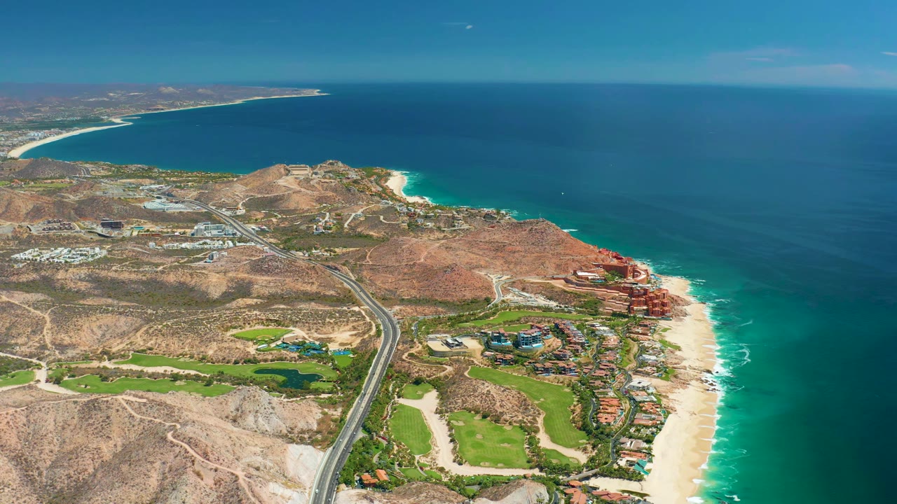 Aerial frame over Marquis Los Cabos showing the highway running toward the shoreline, tan hills, small green fairways, clustered coastal buildings, blue water, and no visible people.