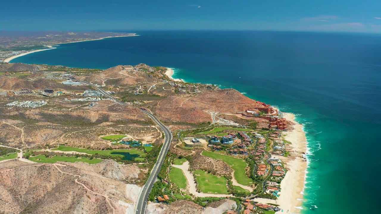 Aerial view over Marquis Los Cabos where the curving coastal highway crosses the lower frame, golf greens sit near the beach, surf traces the sand, and no visible people appear.