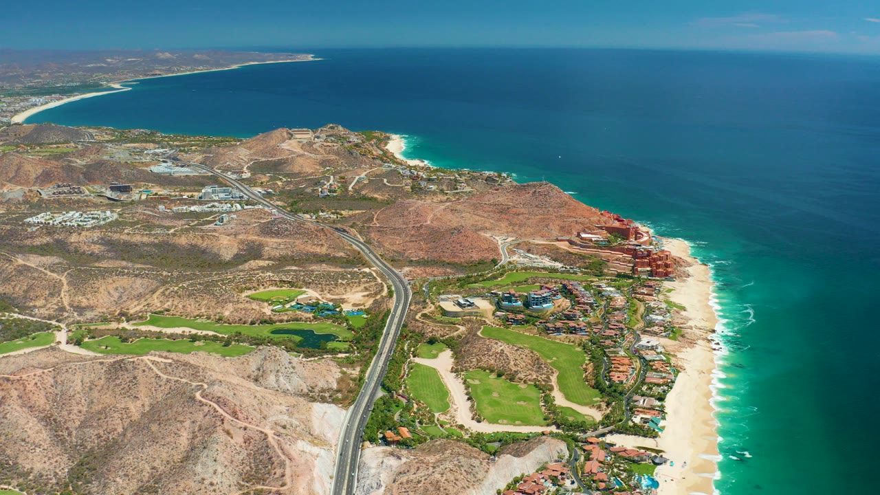 Opening aerial frame over Marquis Los Cabos, Mexico, with a coastal highway running through dry hills toward golf course greenery, sandy beach, turquoise surf, and deep blue ocean, no visible people.