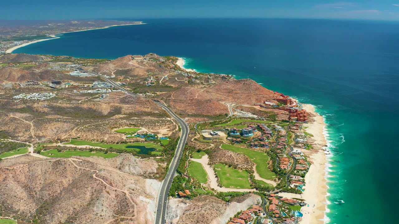 Wide aerial coastline with highway, beach, arid hills, green golf areas, and ocean surf.