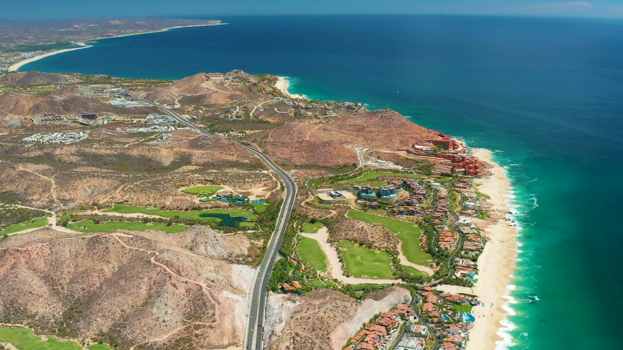 High aerial view over Marquis Los Cabos, Mexico, showing a highway through dry hills, green landscaped areas, beachfront buildings, sandy beach, blue ocean, and no visible people.
