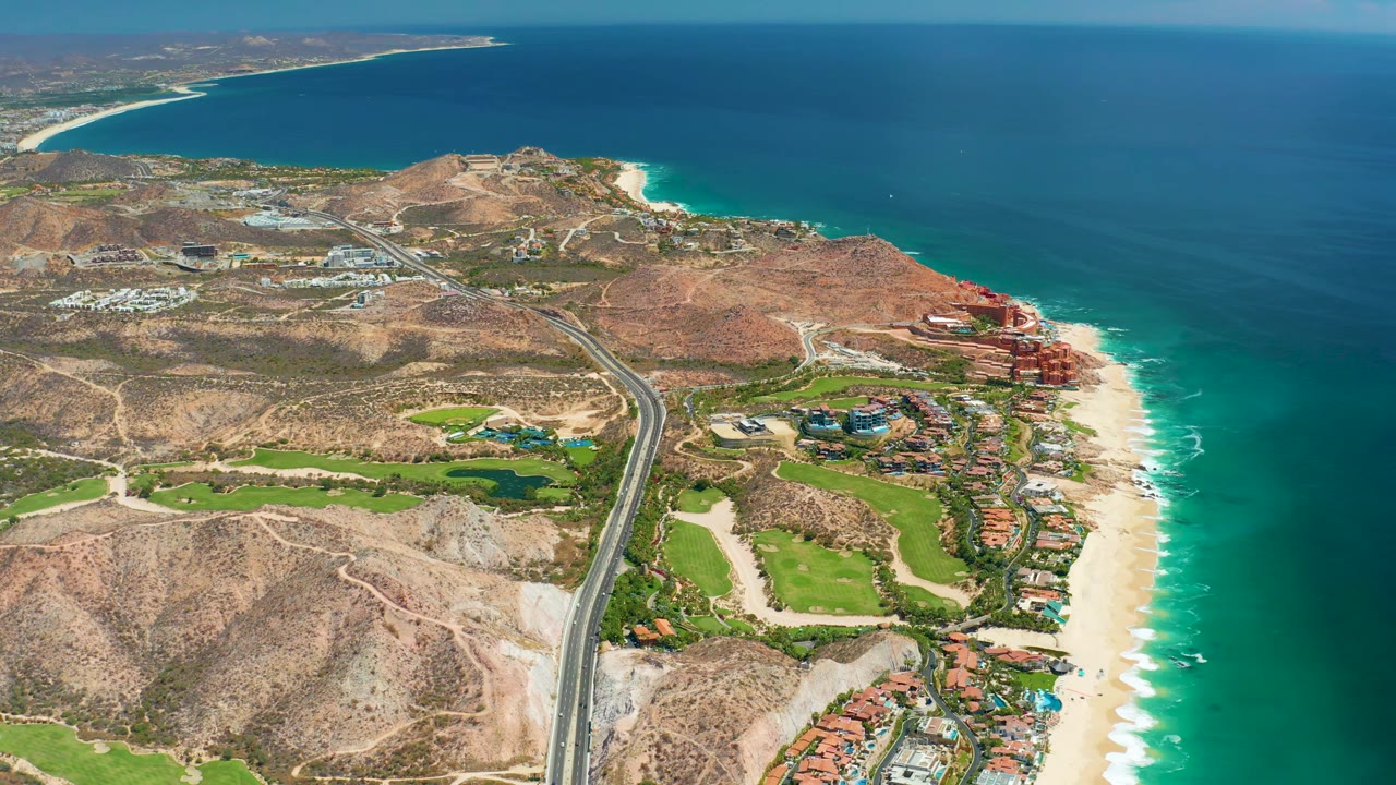 Wide aerial view at Marquis Los Cabos showing the arid hillside foreground, a curving coastal road, green landscaping, shoreline buildings, bright sand, clear blue water, and no visible people.