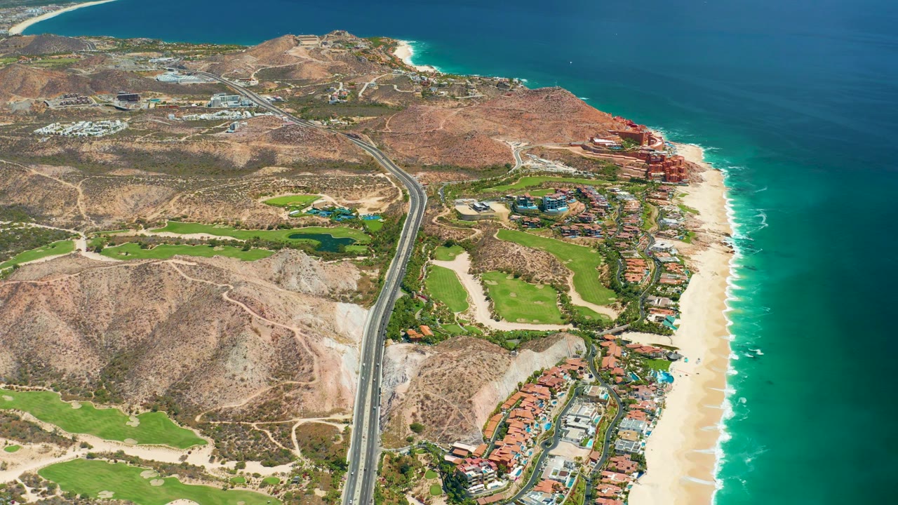 Drone-like view of highway through dry hills near golf greens and sandy coastline.