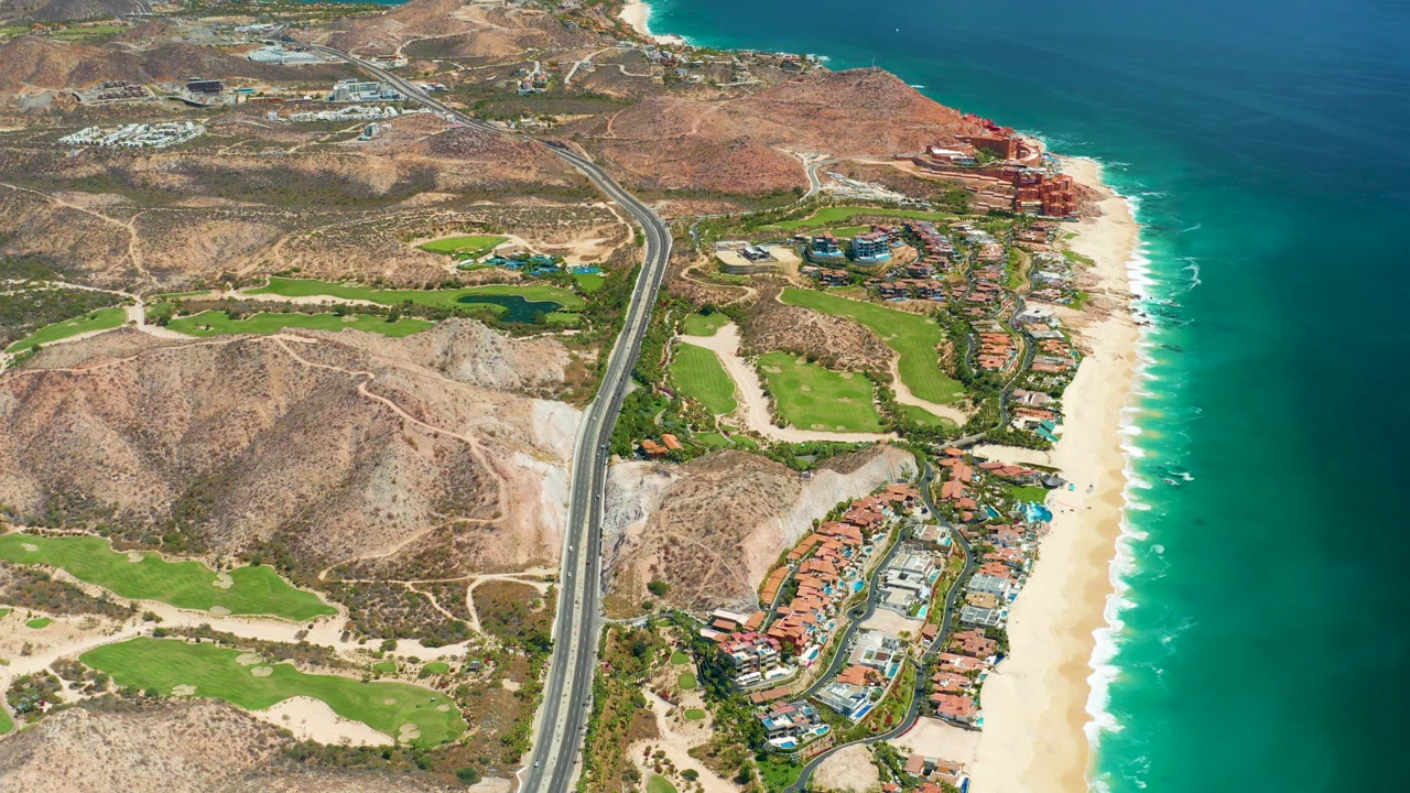 Opening aerial frame over Marquis Los Cabos, Mexico, showing a highway between arid hills, green golf fairways, sandy beach, turquoise ocean, and no visible people.