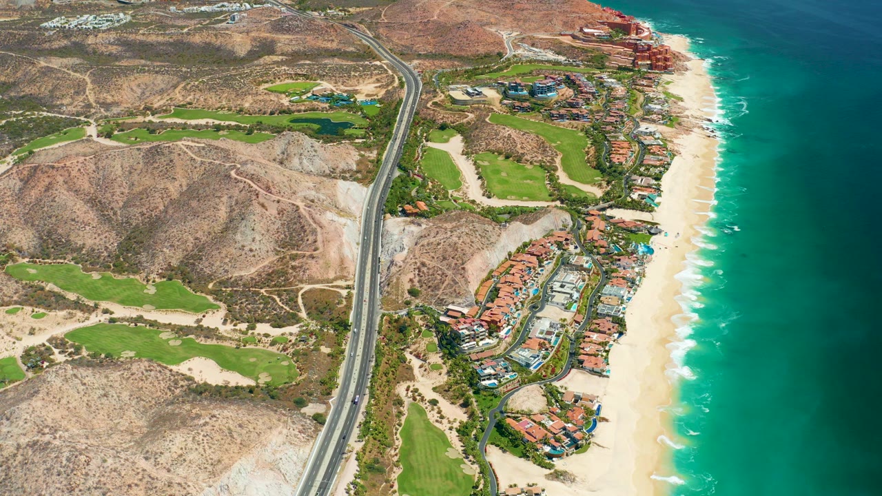 Aerial view over Marquis Los Cabos, Mexico, with a coastal road running past tan hills, green golf areas, resort buildings, pale sand, turquoise ocean, and no visible people.