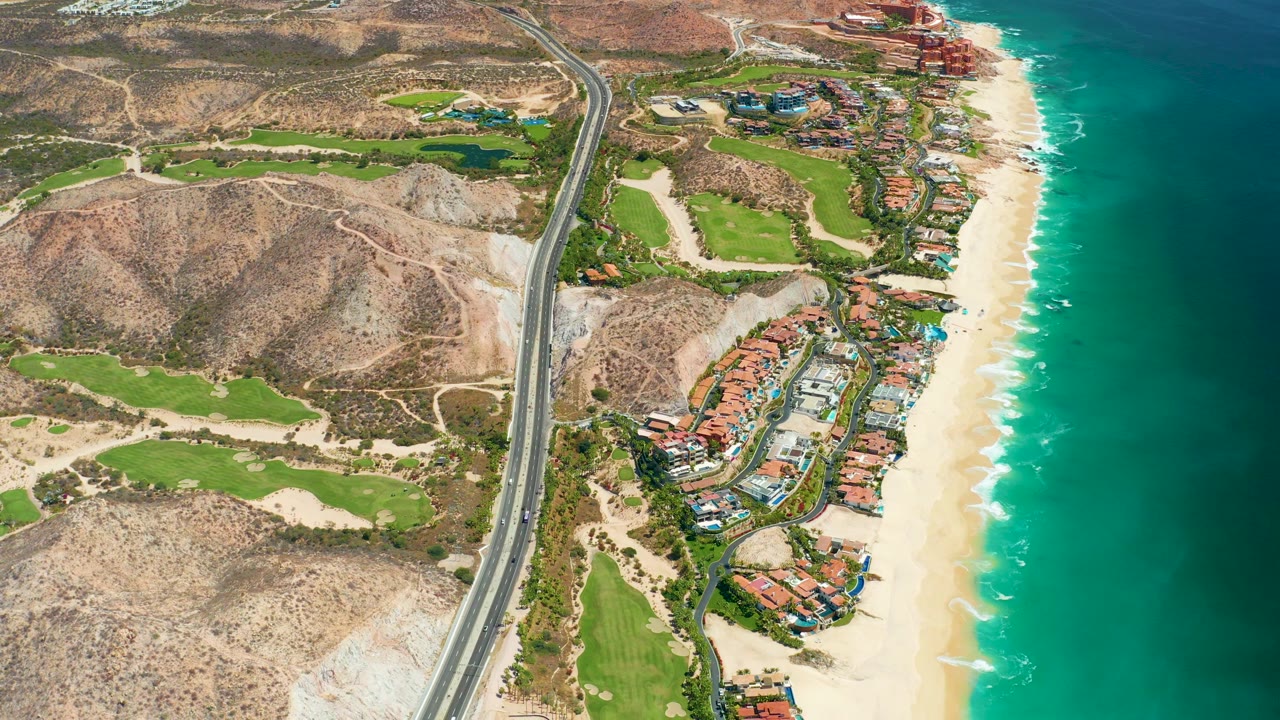 Drone view of coastal road, hillside terrain, buildings, beach, and surf.